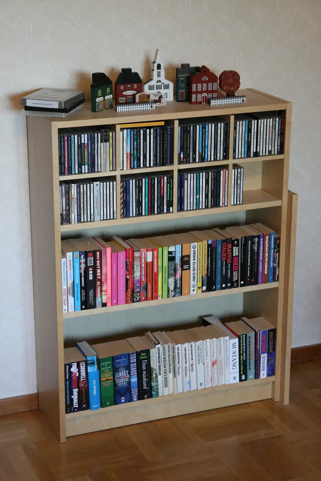 a light wood flat-pack style bookcase full of books and CDs standing against a wall on a wood floor