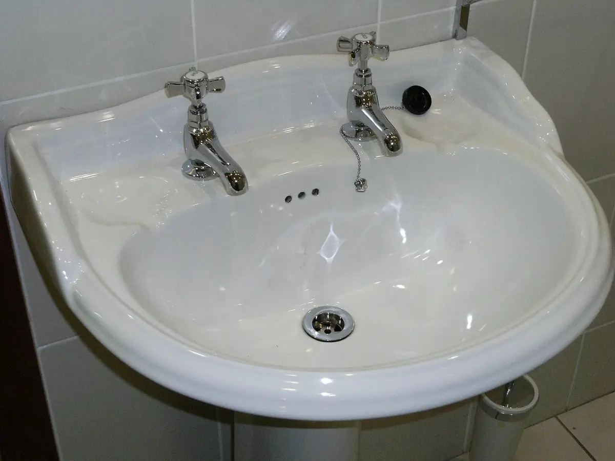 a white bathroom wash basin with separate hot and cold chrome faucets, against white tile