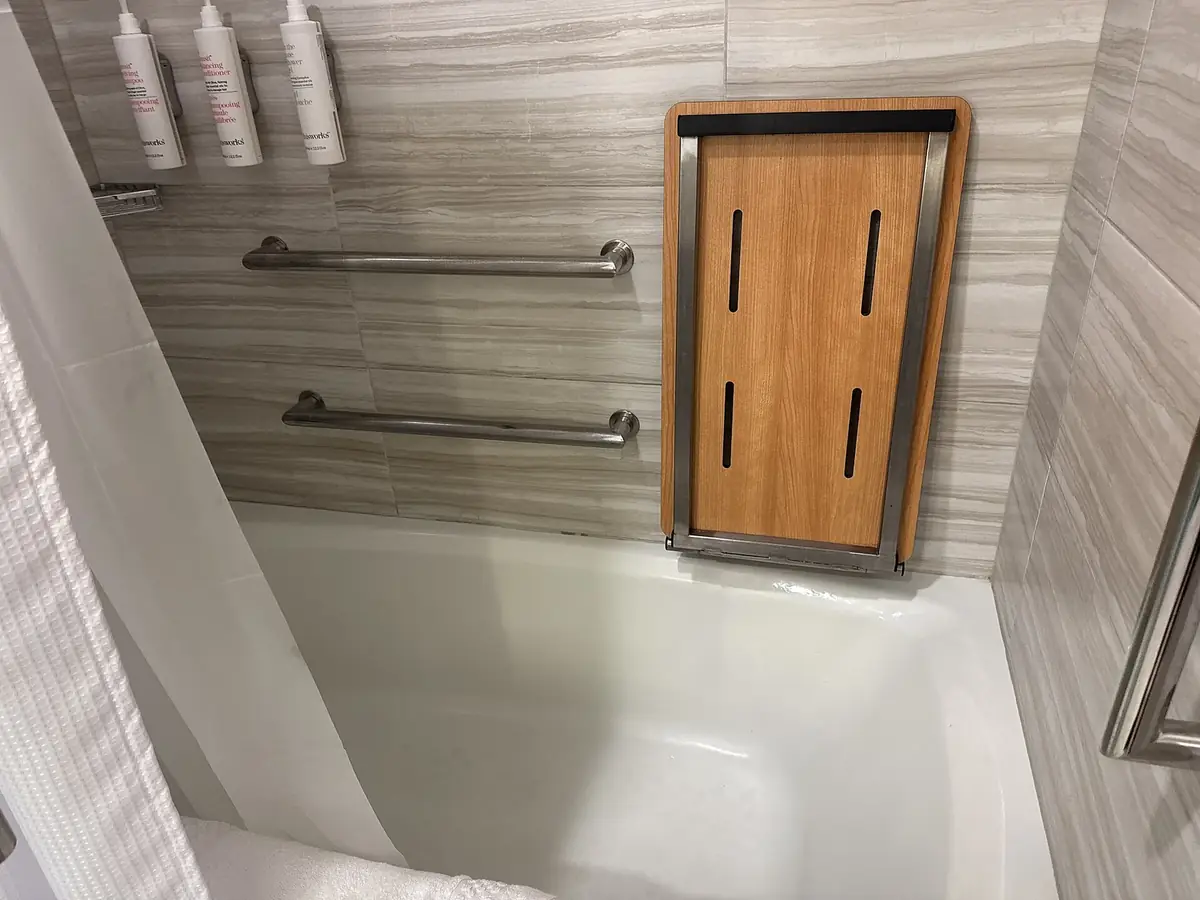 a bathtub with two horizontal stainless grab bars and a fold-down wooden shower seat on a gray tiled wall