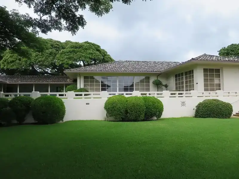 a long single-story white house with a tiled hip roof and a low wall, behind a green lawn and trimmed shrubs in Honolulu