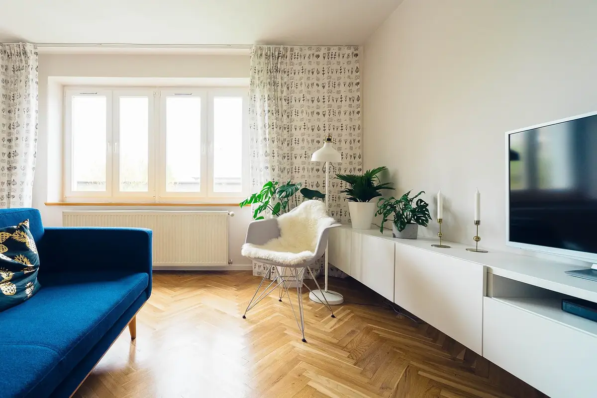 a bright living room with a blue sofa, plants and a flat-screen TV above a long white media console