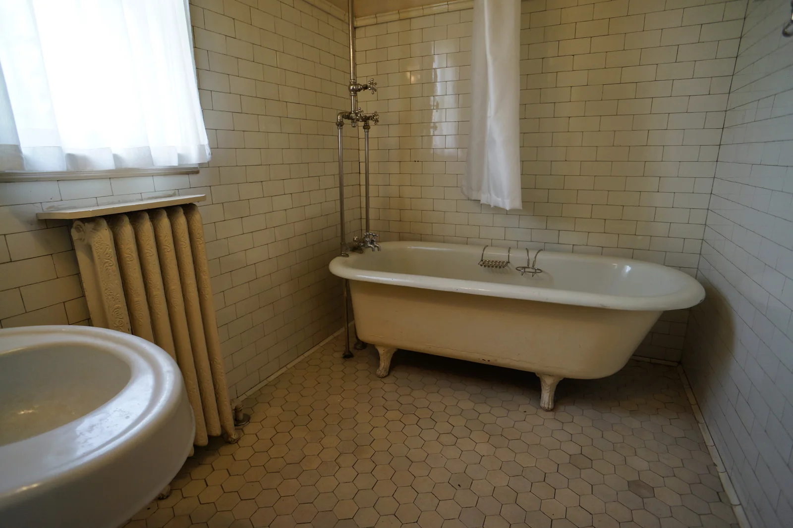 a white subway-tiled bathroom with a clawfoot tub, hexagon floor tile, a radiator and a pedestal sink in the foreground