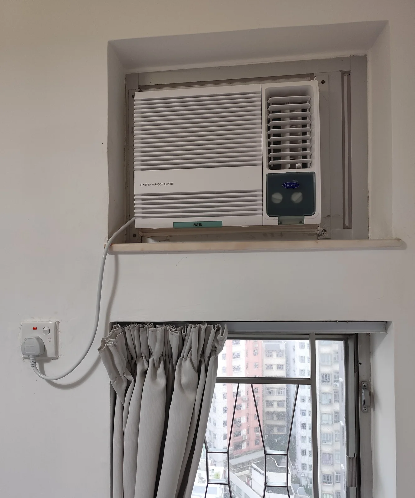 a white window-type air conditioner set into a wall opening above a curtained window, with its power cord running to an outlet