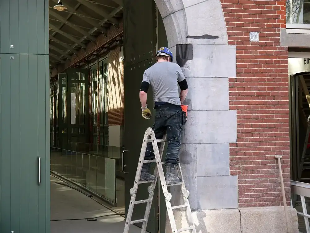 a worker in a hard hat standing on a step ladder, working on the stone frame of a large doorway in a brick building